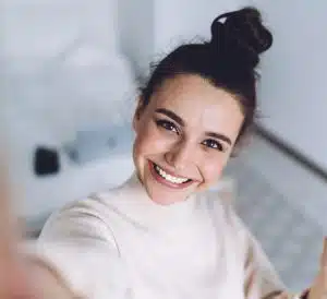 Pretty young female with big smile standing at bedroom after work with laptop and having fun taking light cheerful selfie on blurred background